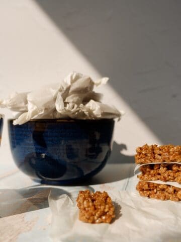 blue bowl full of wrapped grape nuts pasteli. 4 unwrapped outside the bowl next to an espresso.