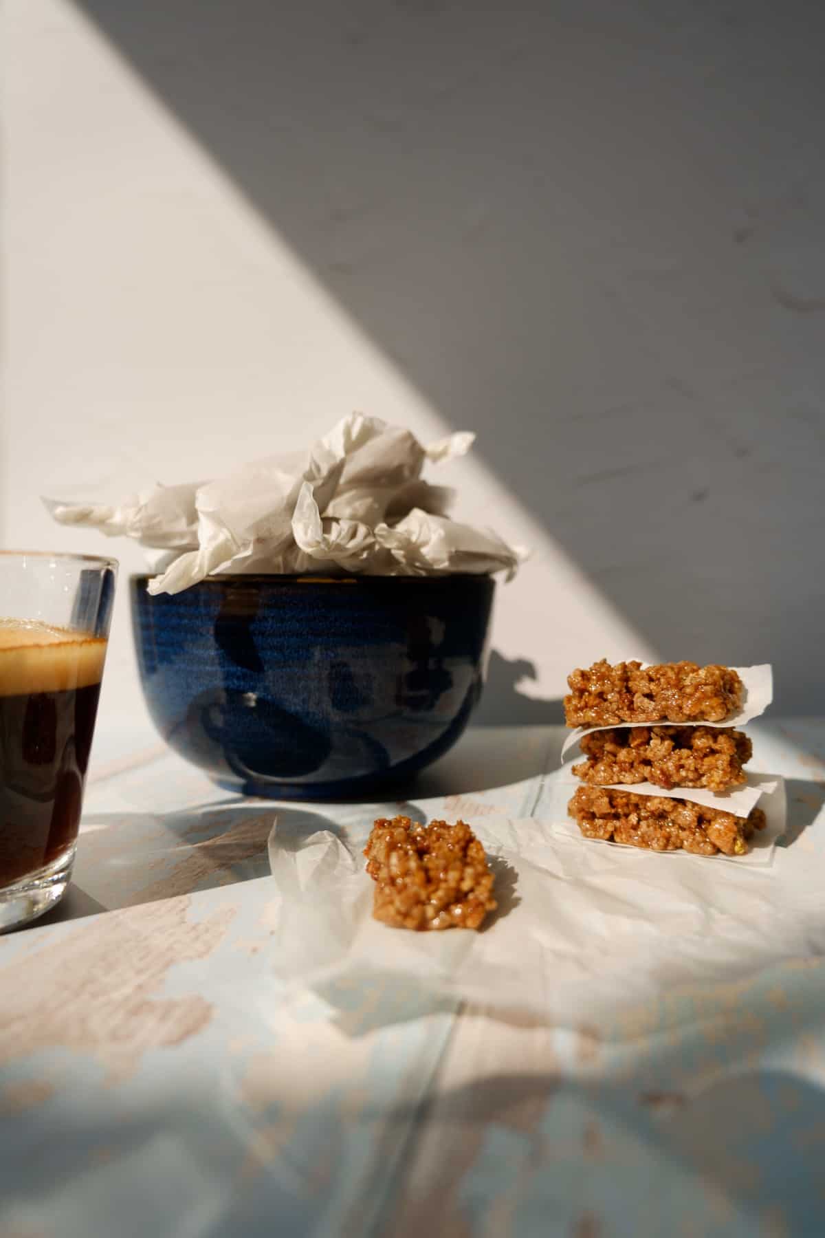 blue bowl full of wrapped grape nuts pasteli. 4 unwrapped outside the bowl next to an espresso.