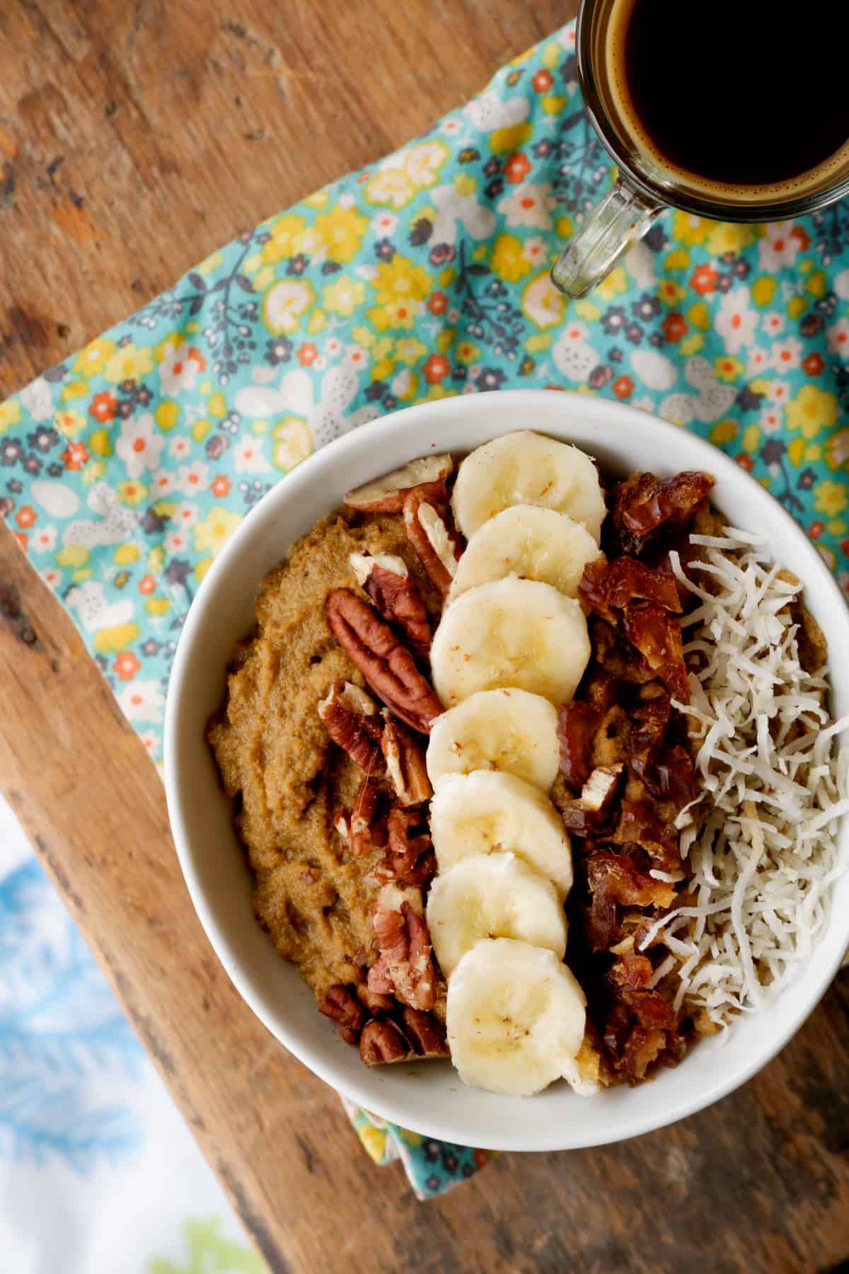 Whole30 sweet potato breakfast bowl close up. On bed tray with a cup of coffee. Topped with bananas, coconut flakes, dates and pecans.